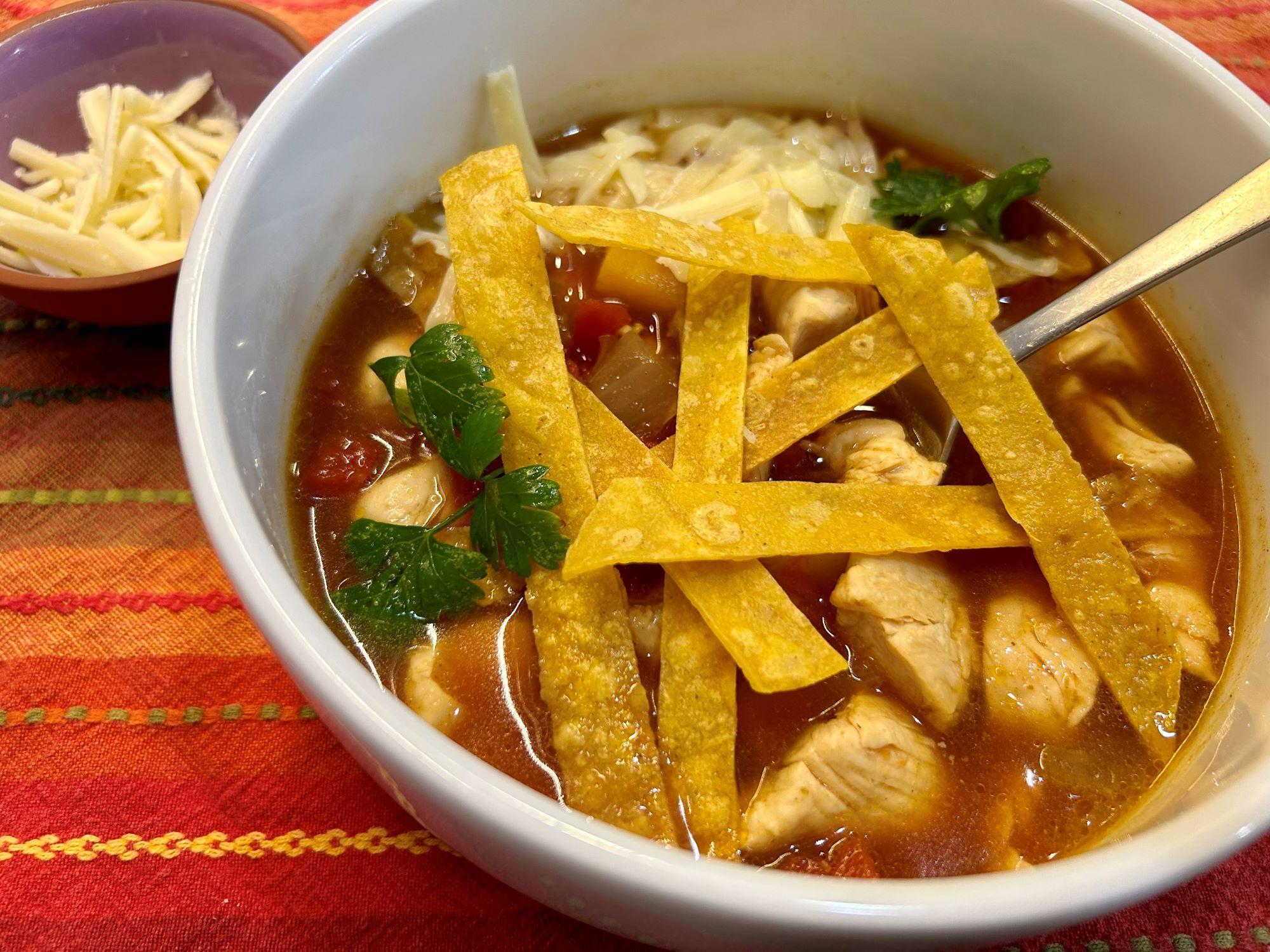 A bowl of chicken and vegetable soup with wonton strips on the top and shredded cheese to the side.