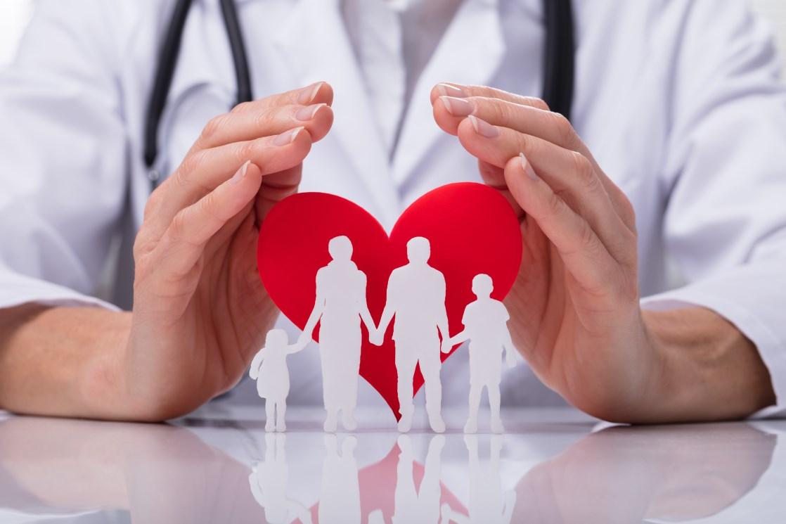 In the image, a medical doctor's hands are shown holding a red paper heart, with a white paper silhouette of a family holding hands.