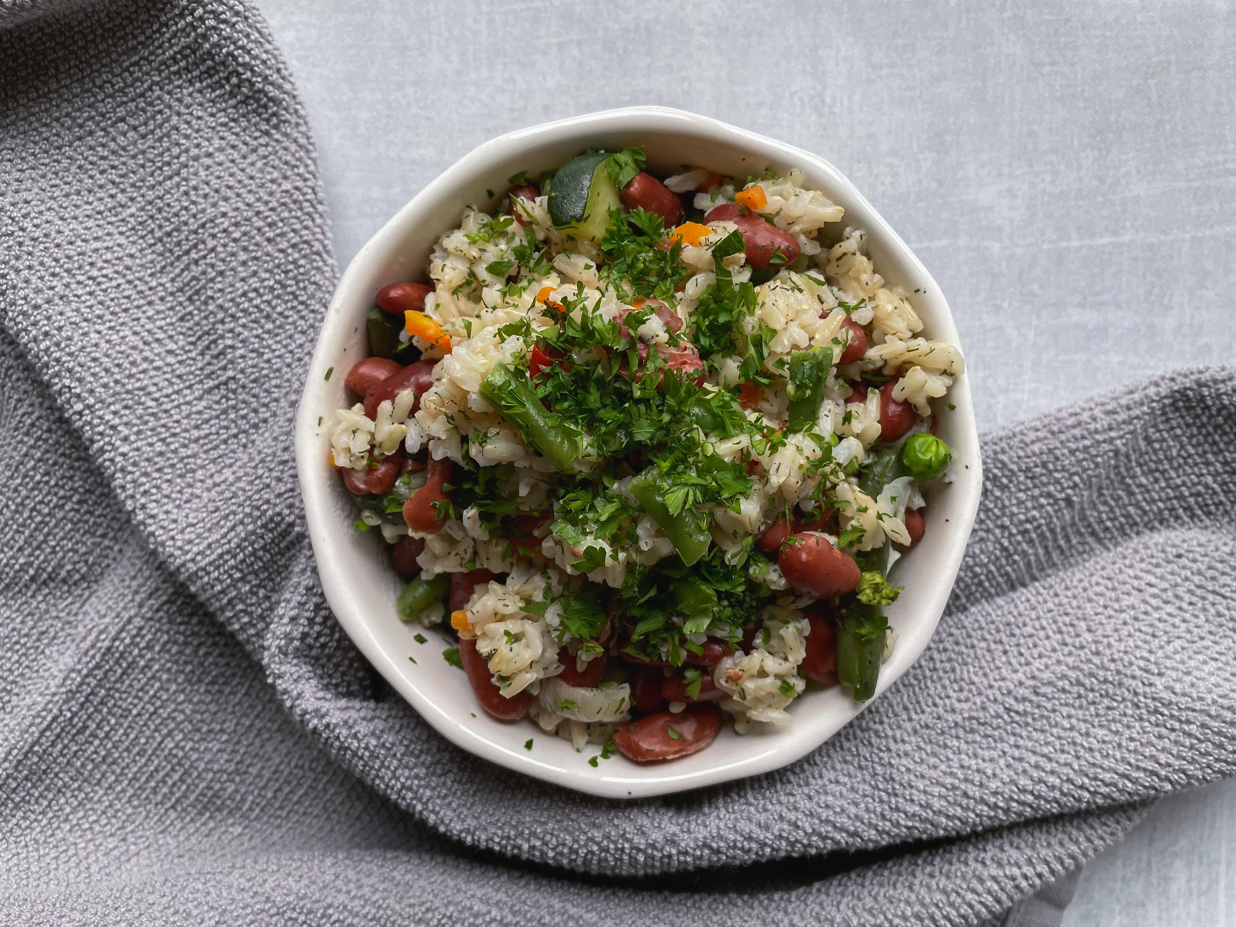 Whole grain rice topped with cooked frozen vegetables and garnished with dried dill.  Rice dish in a white bowl and a gray dish towel on the bottom of image.