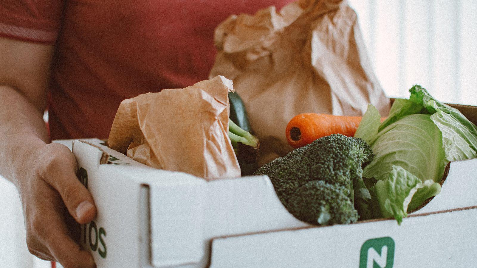 An individual carrying a food produce box with broccoli, cabbage, and carrots in a white box.