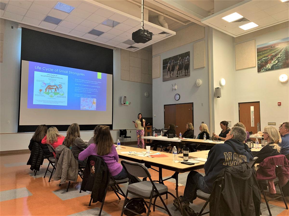 Extension educator is presenting at a workshop with participants seated around tables, watching a PowerPoint presentation.