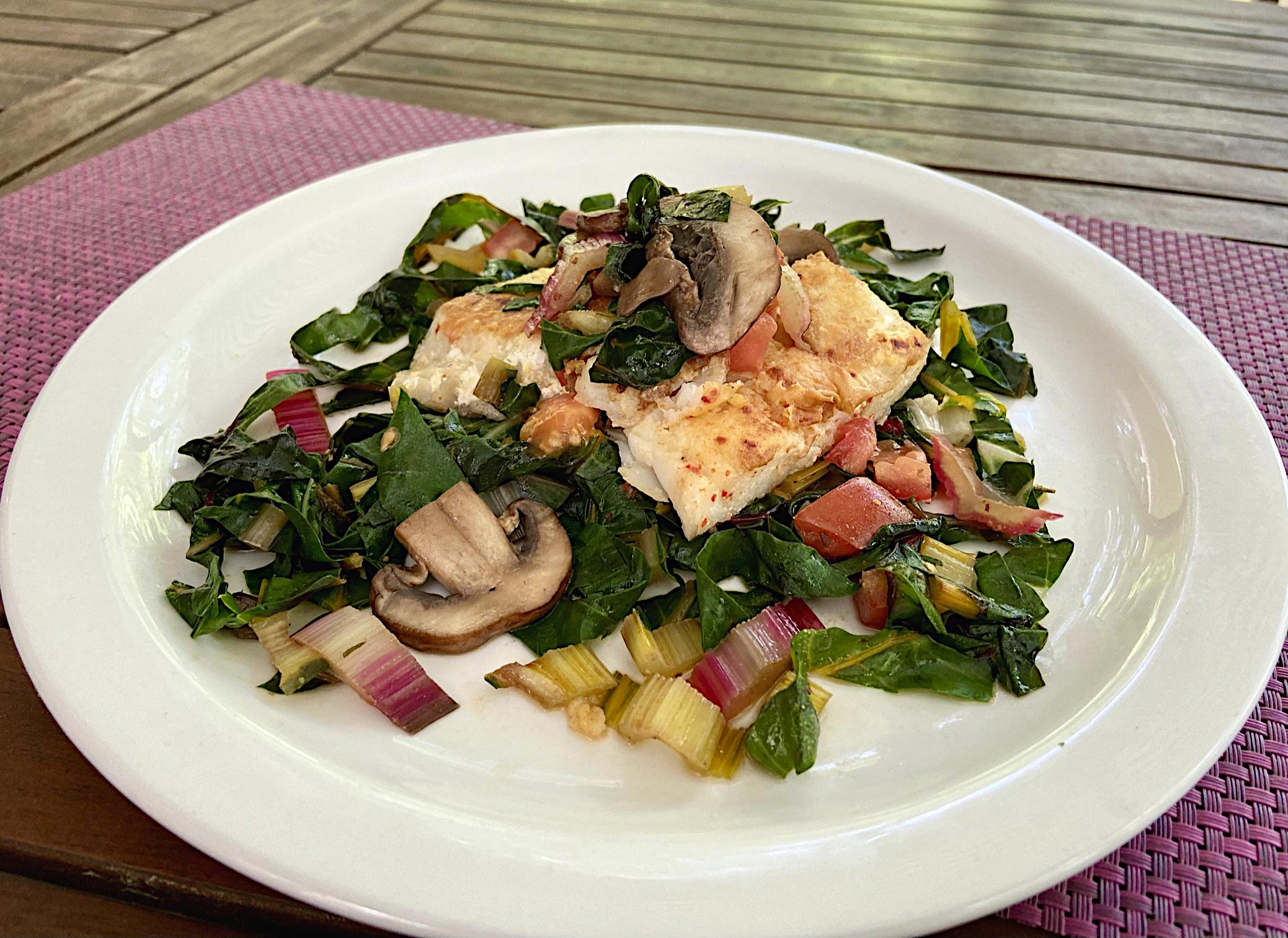 Baked fish on a bed of swiss card and mushrooms lying on a white plate and a pink table mat.